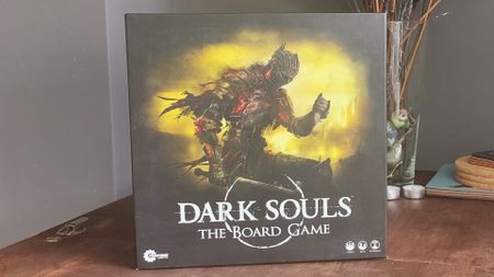 The Dark Souls board game box on a wooden table, with plants in the background