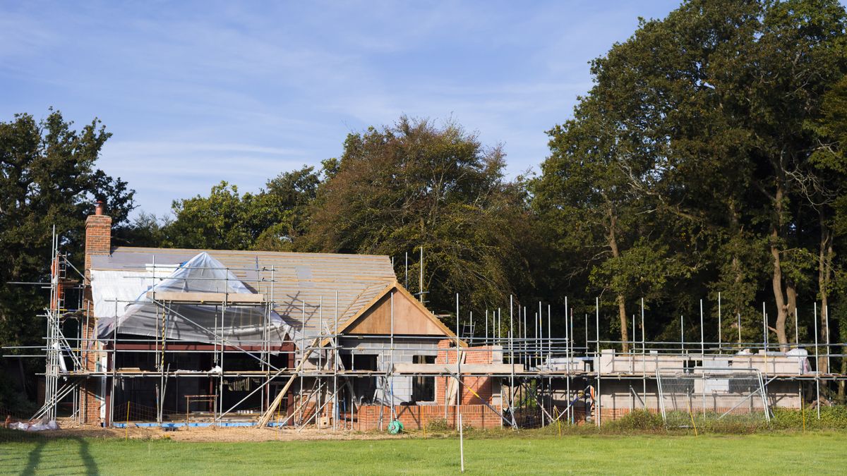 Exterior view of house in the process of being built with scaffolding around it, grass in front and trees behind