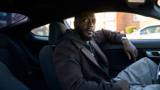 Cross sitting in a car with his arm on the window and looking at something off camera.