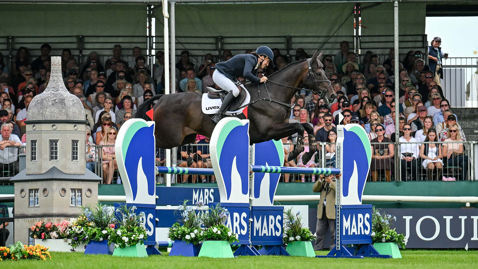 Katie Magee, a rider in a blue coat, steers dark bay horse Treworra over a blue and green showjump at Burghley 2026