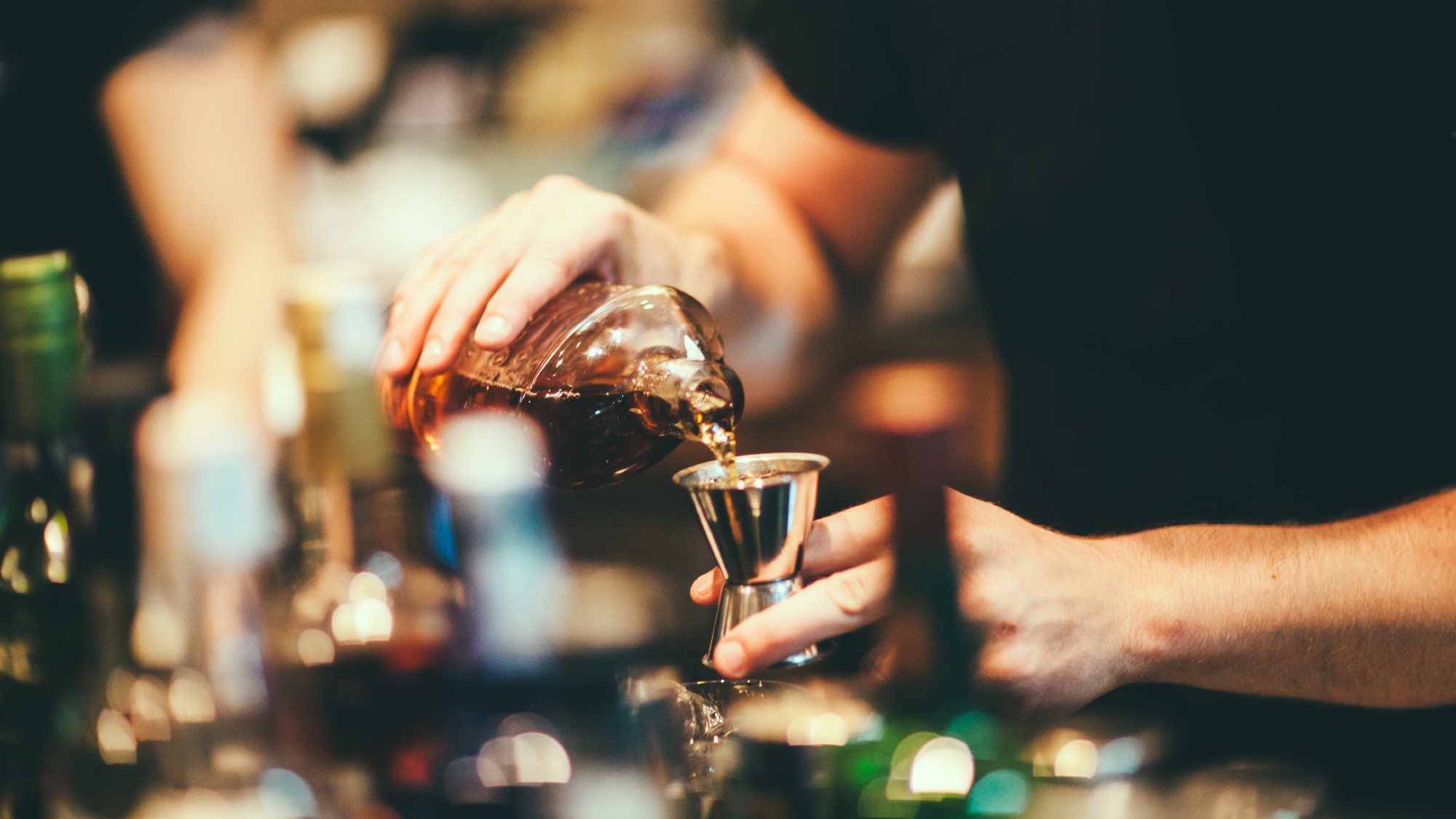a bartender pours brown liquor into a metal jigger