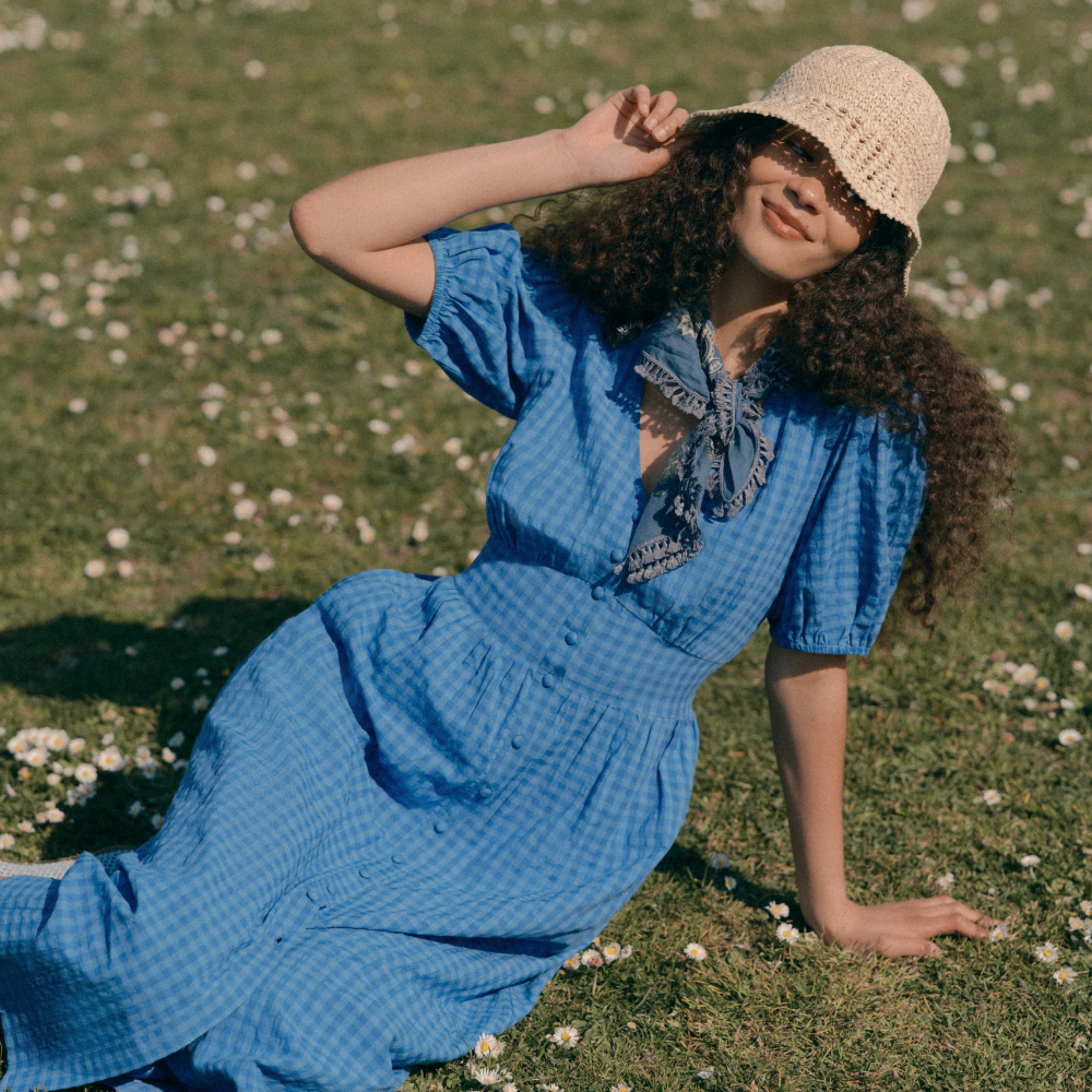 Model sat on grass wearing a blue gingham dress and neck scarf with a beige crochet bucket hat all sold at Finery London