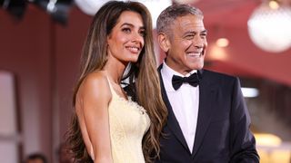 Image of Amal Clooney, wearing a pastel yellow gown, and George Clooney, wearing a black tuxedo, smiling on the red carpet