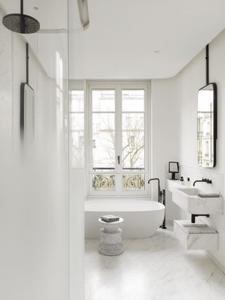 white apartment bathroom with marble floor and wall mounted vanity, mirror, black fixtures and fittings, white tub, shower, stool