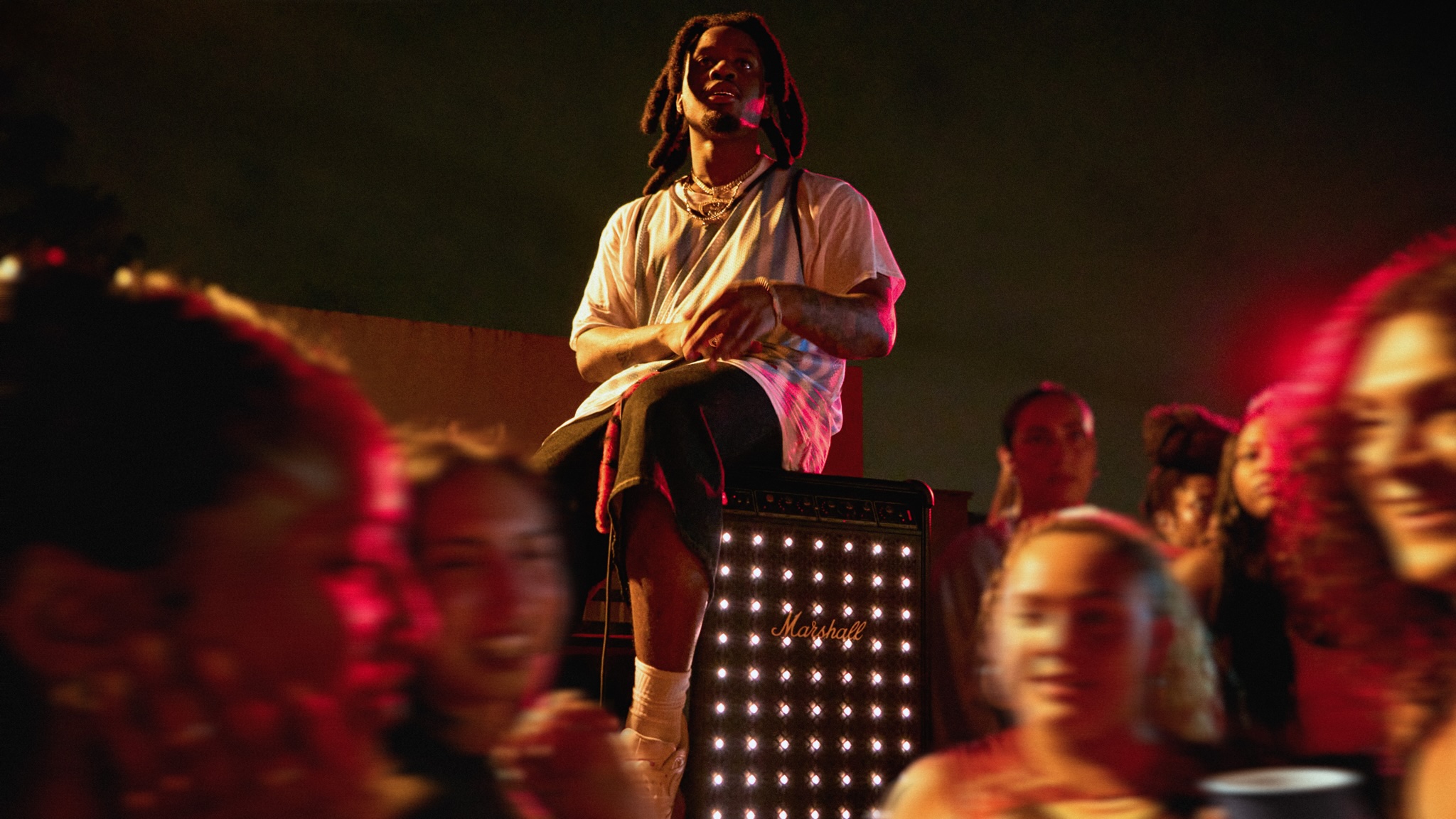 Denzel Curry atop a Marshall Bromley 750 party speaker, at a party. It&#039;s night time and the speaker&#039;s lights are visible on the front.