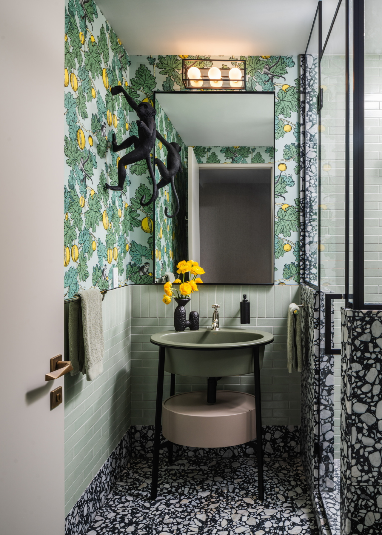 a bathroom with a freestanding sink in two sections in green and pale pink with black legs, pale green tiles with a bright green and yellow patterned wallpaper and a black and white tiled floor and bottom section of the black glass shower screen