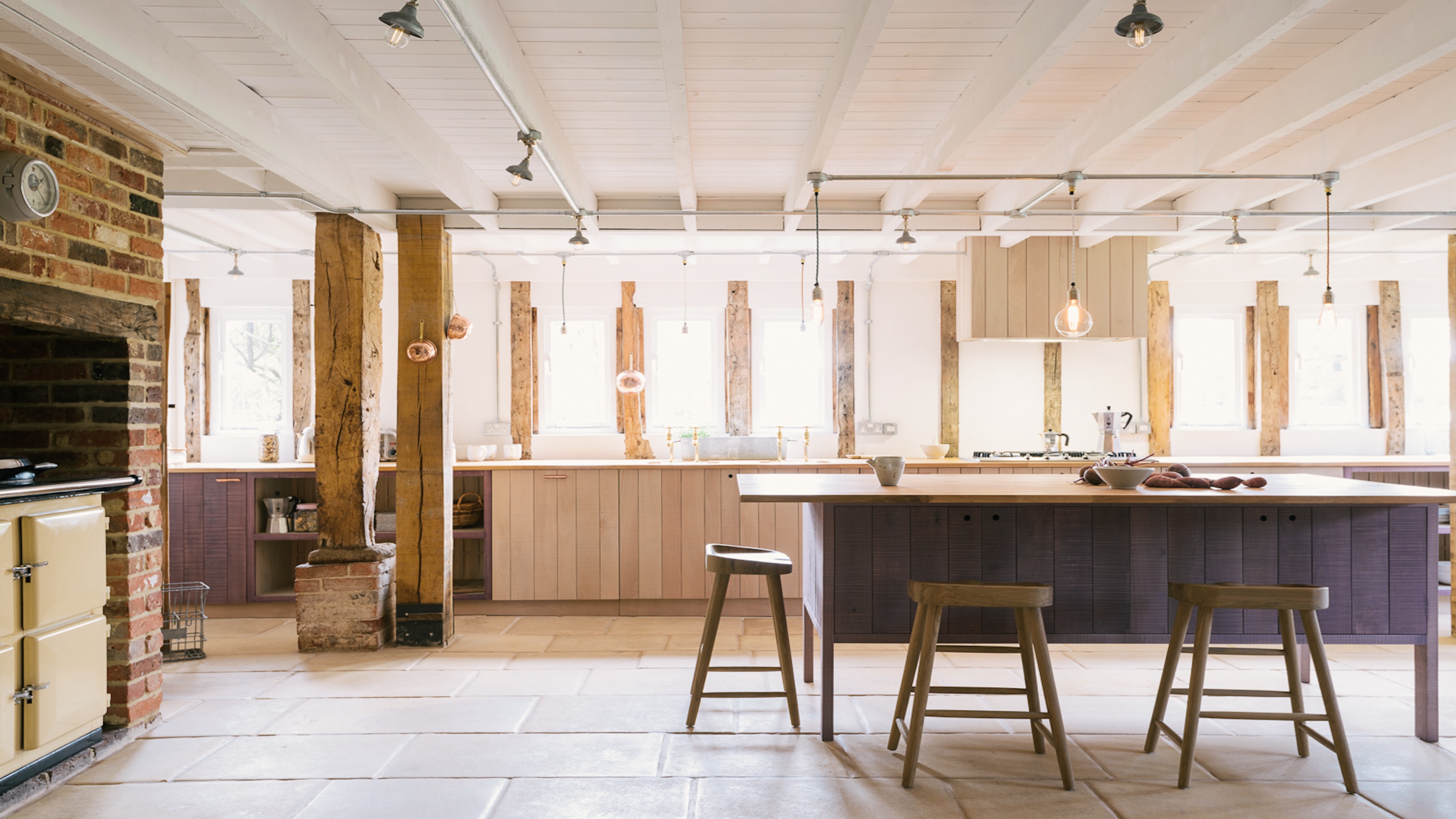 A kitchen with a large island in the middle and bar stools around it as well as a view of the stove by a brick worked wall 