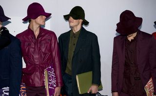 Four male models wearing looks from the Burberry Prorsum collection. One model is wearing a dark blue jacket. Another model is wearing a maroon belted jacked and maroon trousers. The third model is wearing a dark olive green shirt, dark blue trousers and a dark blue jacket. And the fourth model is wearing a dark coloured belted piece with dark purple jacket over the top. All models are wearing hats and holding notebooks
