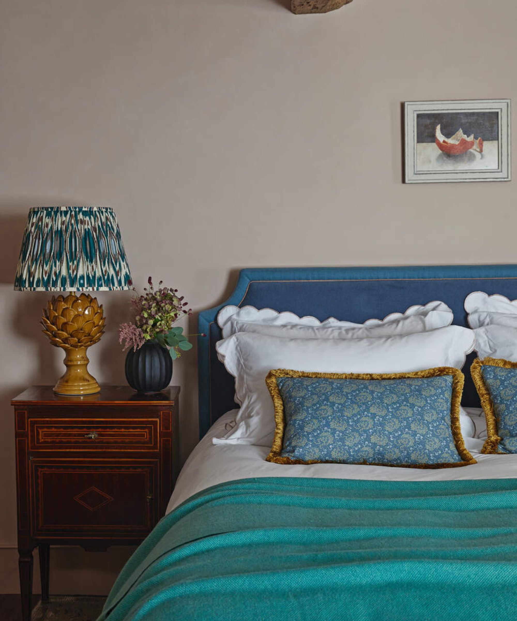 White and blue pillows on a blue bed frame against cream walls; to the left, a lamp on a wooden nightstand.