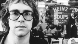 Elton John poses for a portrait wearing glasses in front of a sign that says "Heinz Hot Soups to take away"