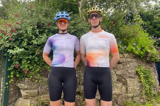 Female and male rider standing in Santini Ombra jerseys infront of a stone wall with planting in the background