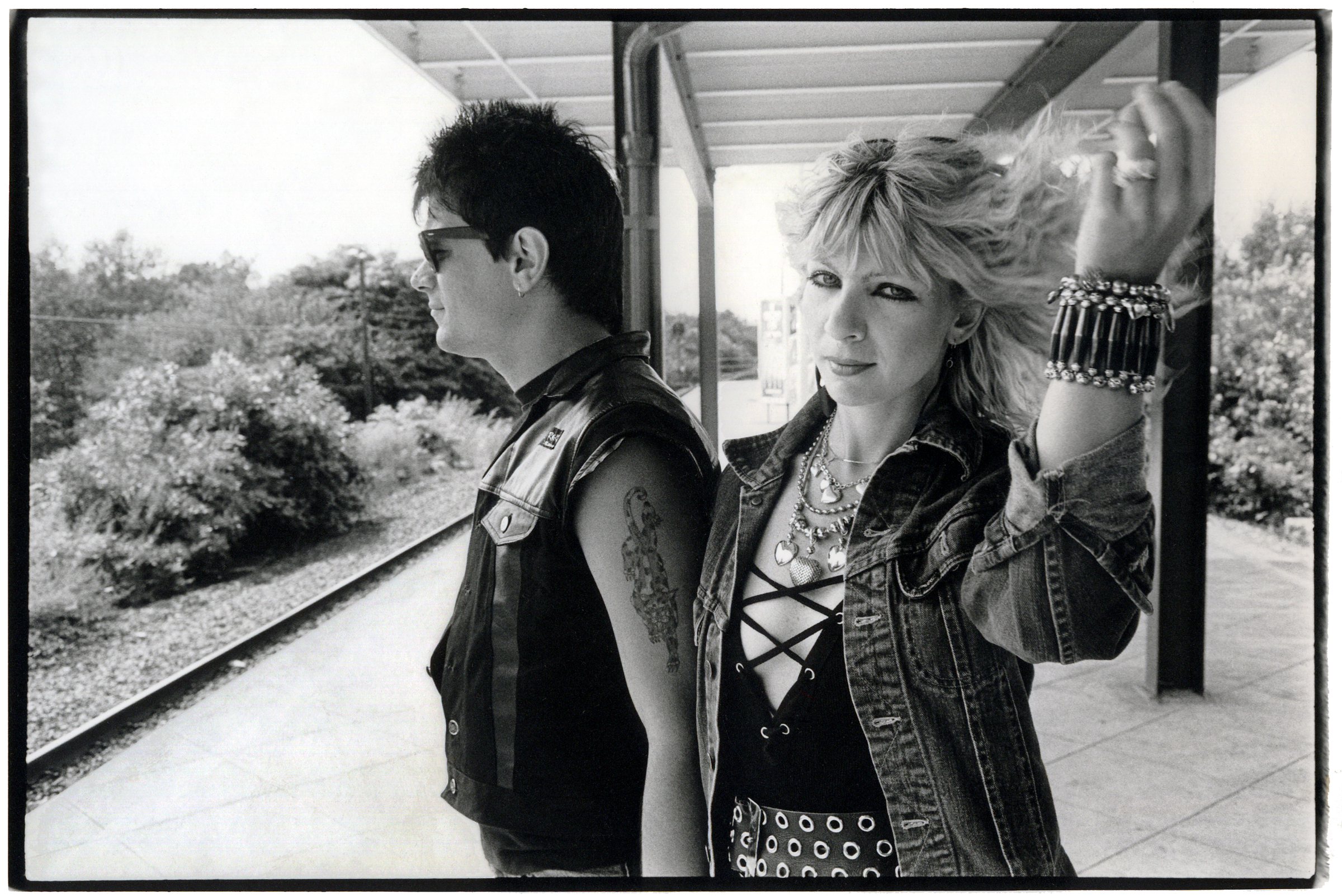 A punk couple stand on a train platform, the man in sunglasses and a leather vest, the woman in a denim jacket and bracelets, holding up her blonde hair