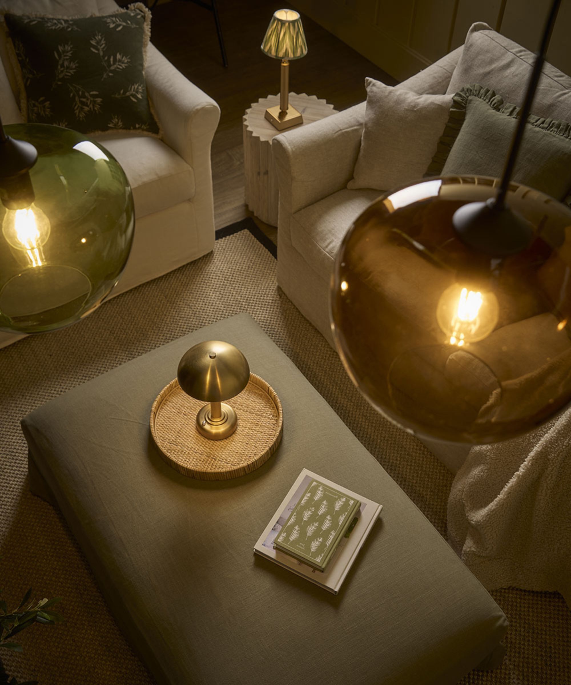 A cosy seating area with cream armchairs and a green fabric ottoman topped with a golden mushroom-shaped lamp. Overhead, amber-toned glass pendant lights illuminate the room, which is decorated with natural textures and earthy green accents.