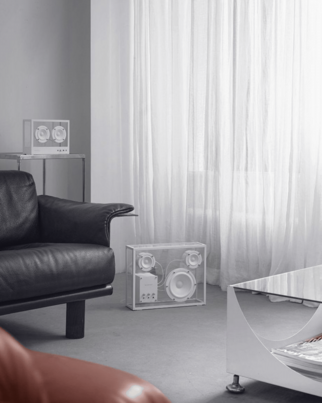 A transparent speaker with white detailing, shown in a rotating gif as styled in a minimalist, monochromatic living room and as a product against a pale gray background.