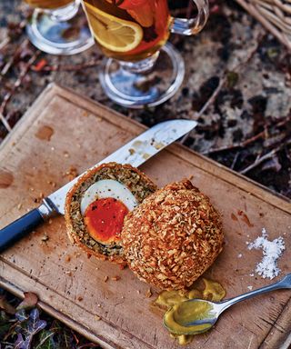 a chopping board with a scotch egg sliced in half with a teaspoon with mustard