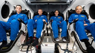 four astronauts in flight suits seated inside of a spacecraft