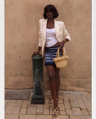 Woman wears linen blazer, white tank top, crotchet blue miniskirt and raffia bag with matching sandals standing on street.
