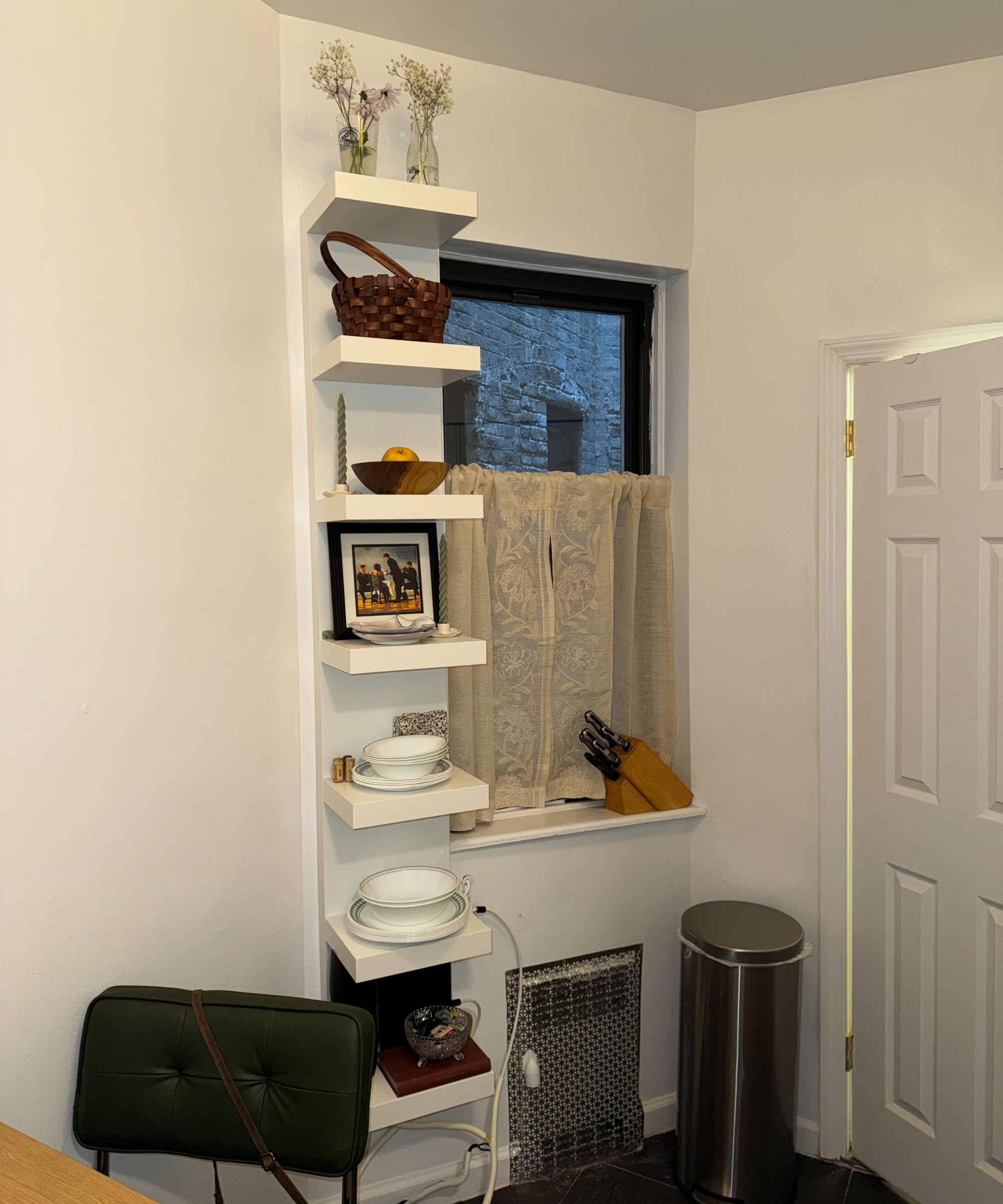 a tall white shelving unit in a kitchen next to a window with cafe curtains hung across