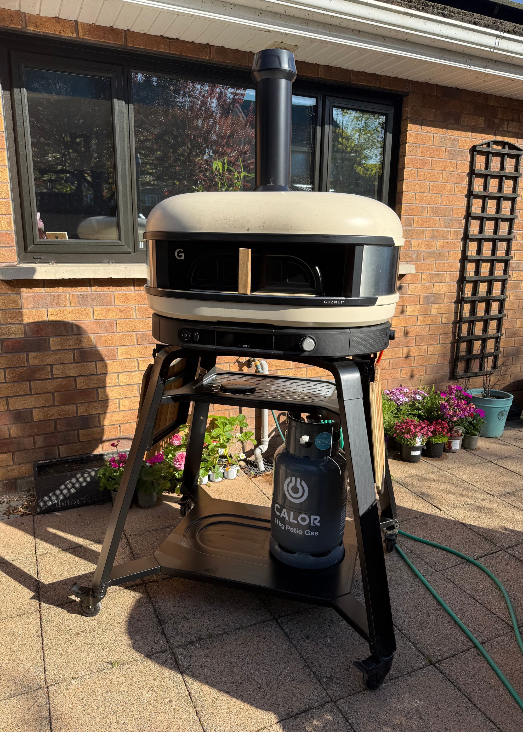 Gozney's Dome XL gen 2 pizza oven in front of a brick house with black-framed windows and plants running along the front