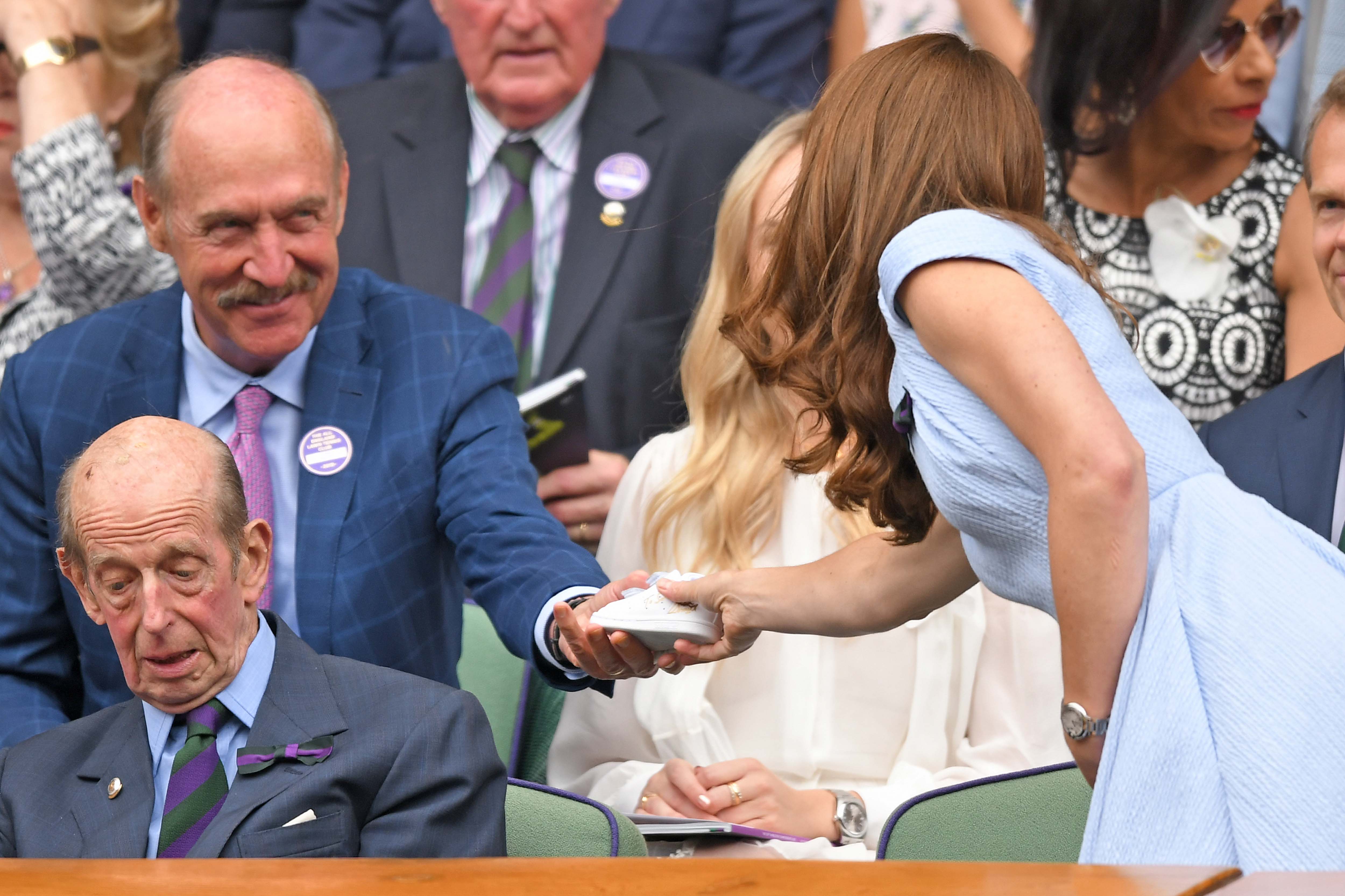 Stan Smith, wearing a navy suit, hands a white Adidas Stan Smith sneaker to Kate Middleton, which he has signed for Prince Louis in gold ink