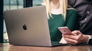 Two people standing around a MacBook. The person on the right of the image is holding an iPhone in their left hand