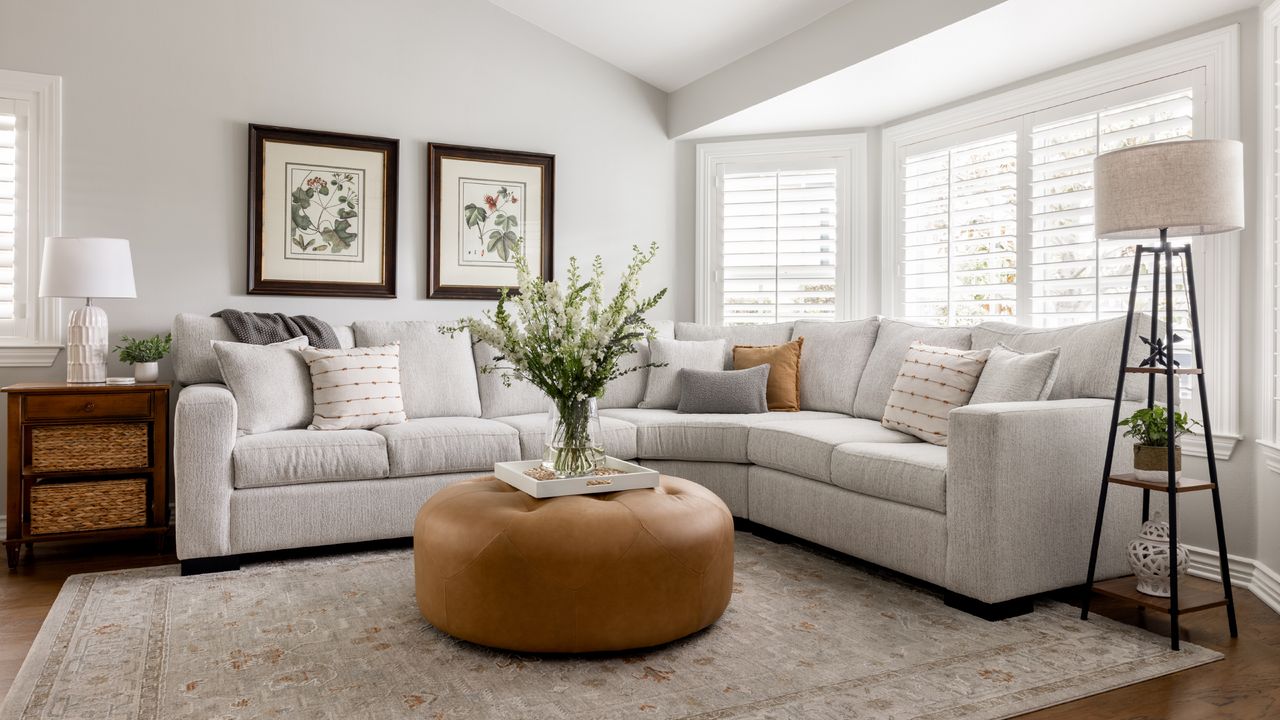 living room with white walls, shutters, pale gray sectional, floor lamp with storage,tan leather footstool, artwork, vase of flowers, neutral scheme