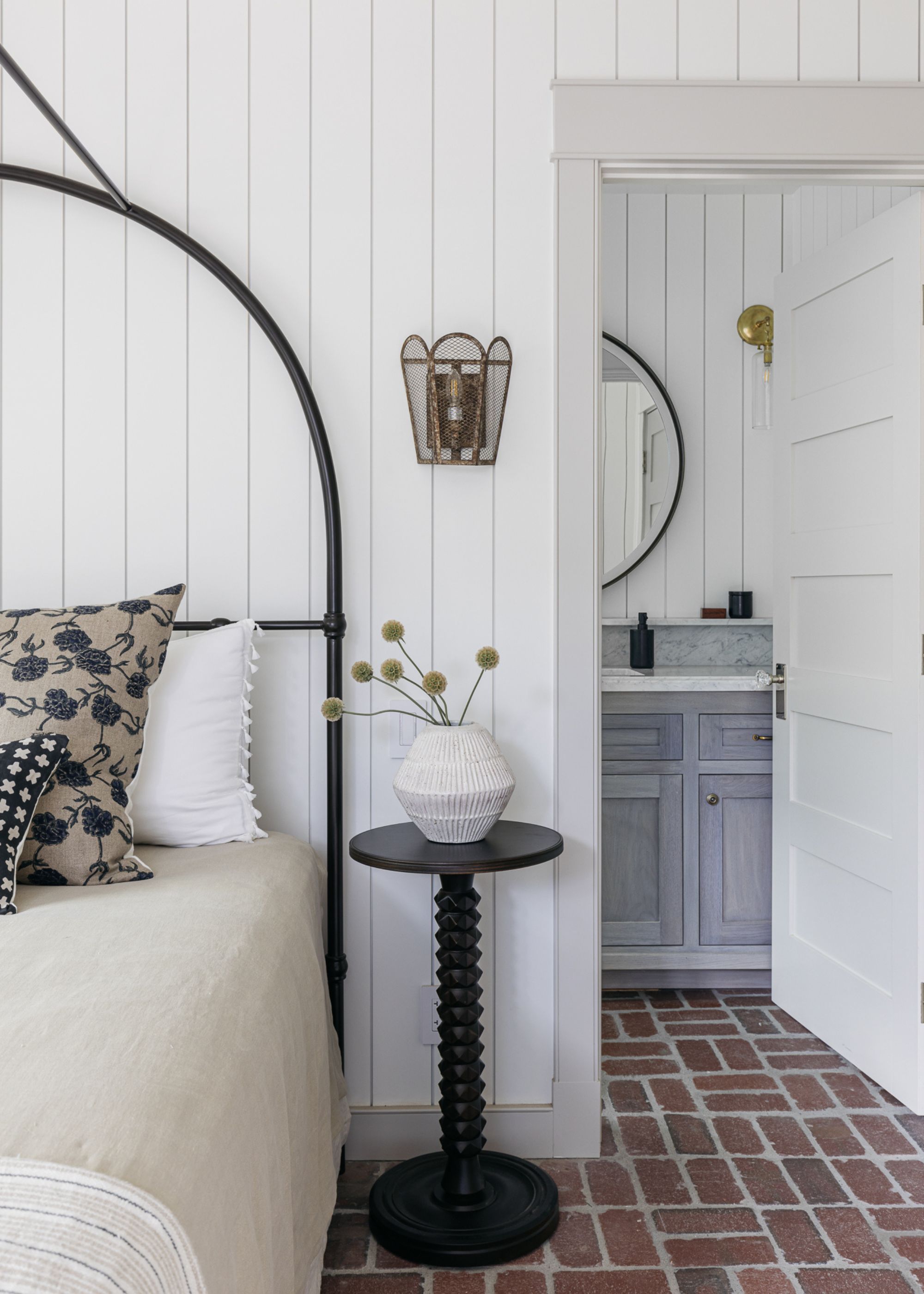 A white coastal bedroom with white paneling walls, neutral bedding, brick floors, and a door leading into a bathroom.