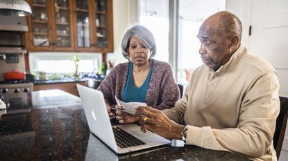 A retired couple sit at their kitchen island and look at paperwork and a laptop.