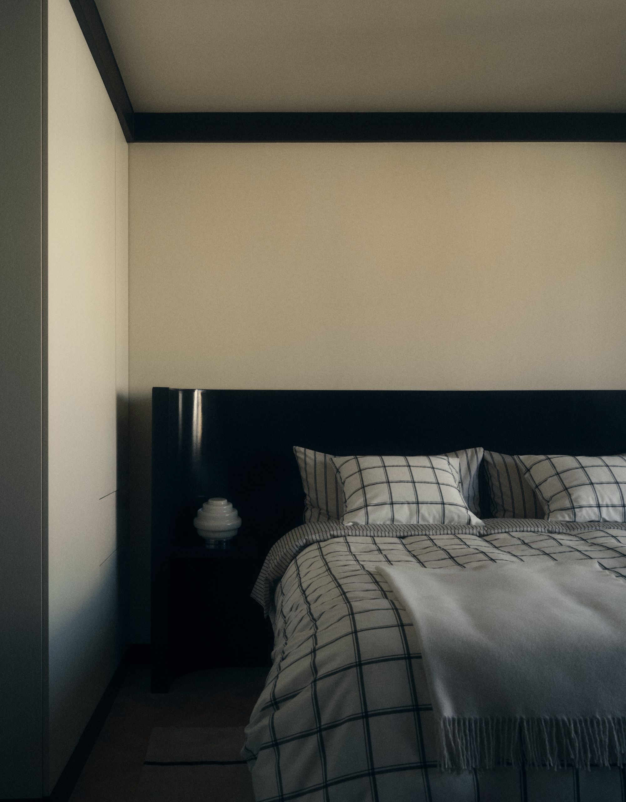 A minimalist bedroom with cream walls and an extended black curved edge headboard in a matte sheen with checked black and white bedding with an off white throw on top and a small white bedside lamp