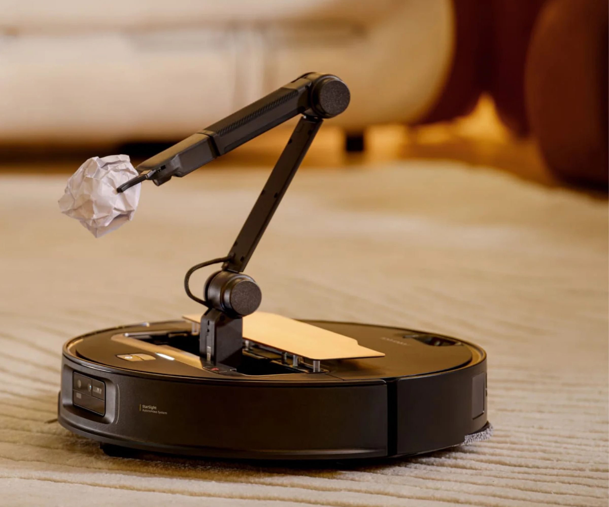 Roborock Saros Z70 robot vacuum with a mechanical arm holding crumpled paper, on beige rug with blurred-out sofa in background.