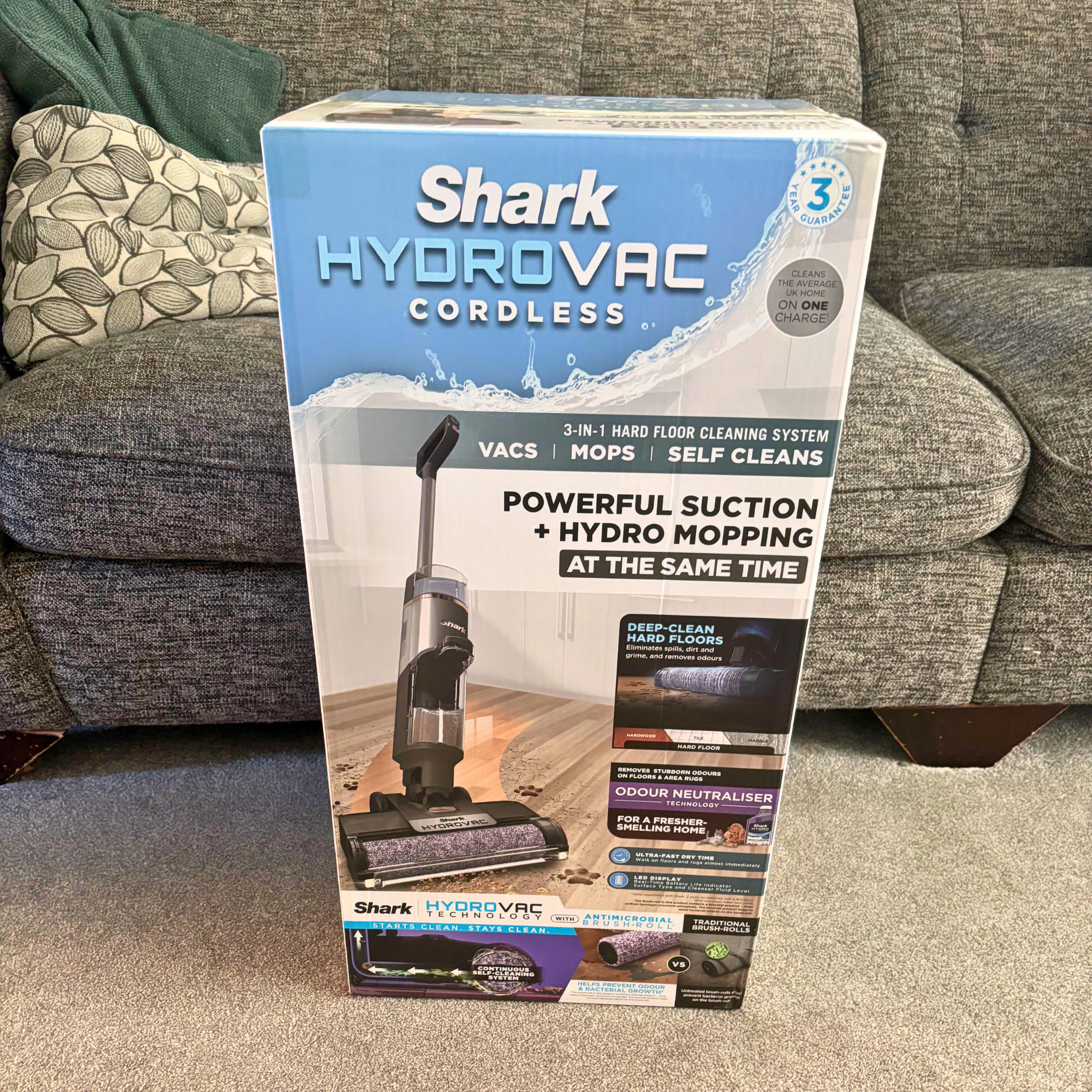 Shark HydroVac Cordless Hard Floor Cleaner  branded box positioned on carpet in front of a grey sofa
