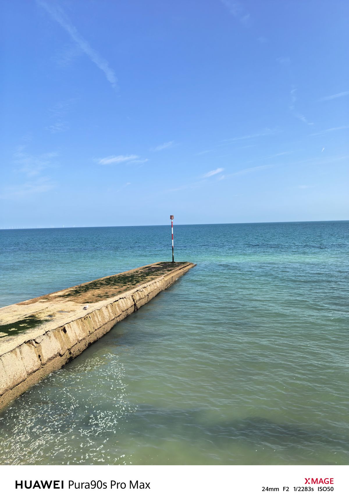 Concrete groyne and red-and-white navigation marker stretching into the sea under a bright blue sky.