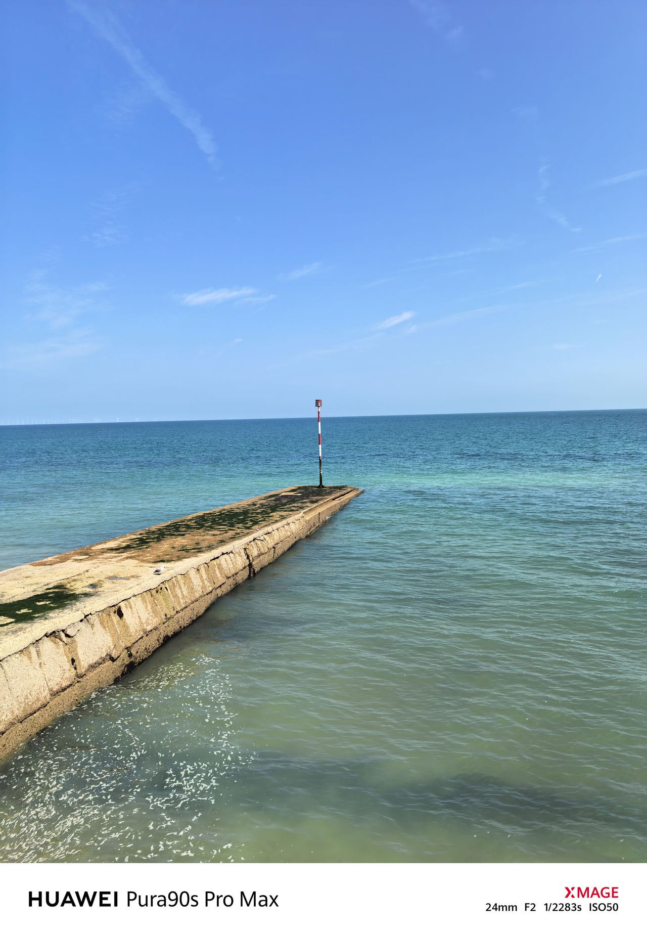 Concrete groyne and red-and-white navigation marker stretching into the sea under a bright blue sky.