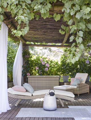 a stunning greek-inspired pergola with soft wood flooring, white linen curtains, and light brown sun younger, chair and ottoman, with a yoga mat and side table with a kettle on