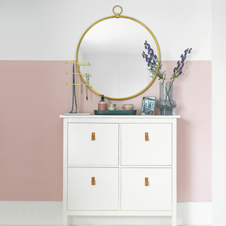 White chest of drawers against white wall with pink painted square, large round mirror on the wall above.