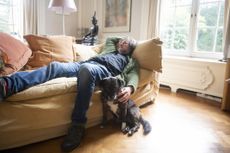 The man is lying on a sofa and the dog is sitting next to him on the floor.