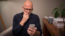 An older man looks uncertain as he looks at his smartphone while sitting next to his desk.