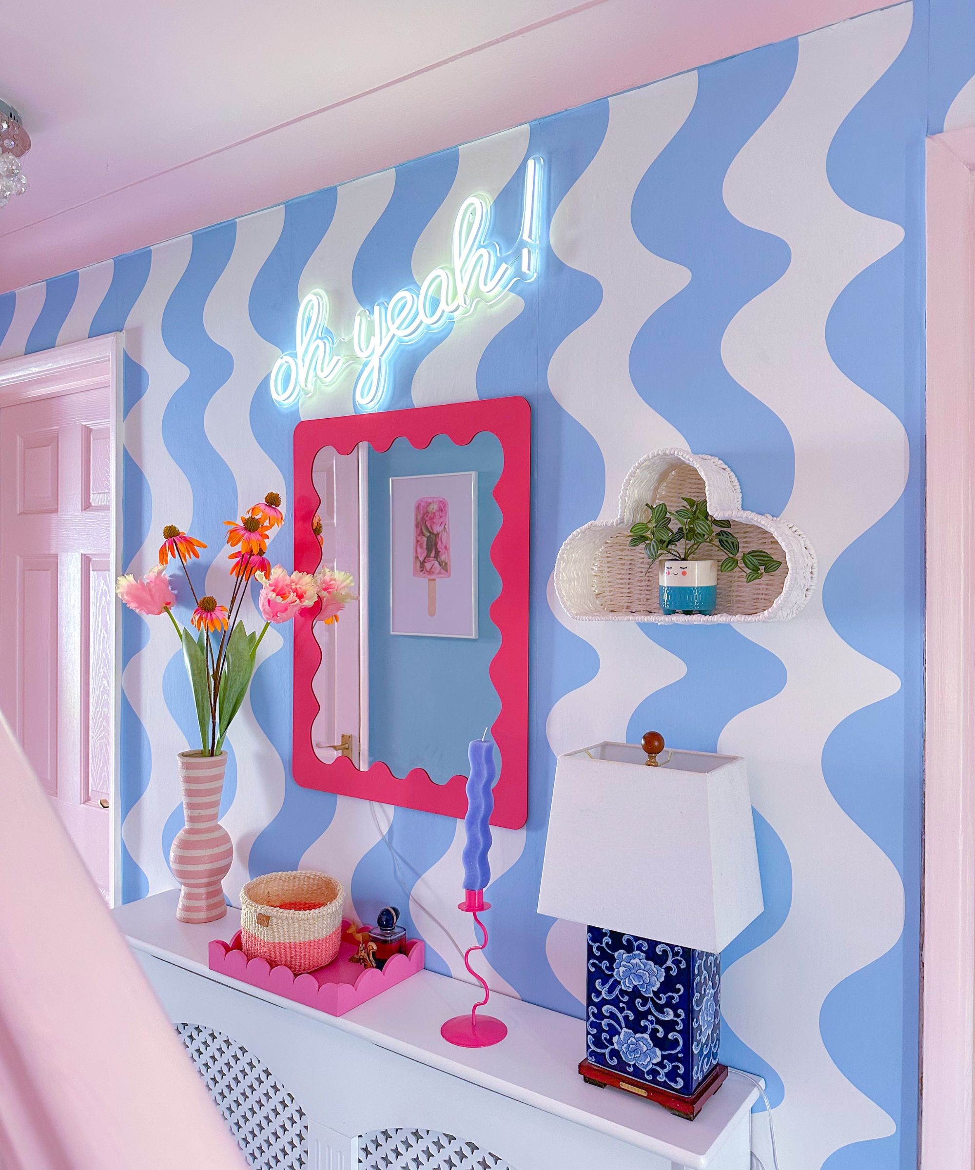 A hallway wall painted with bold blue and white wavy stripes. A hot pink mirror, neon sign saying &amp;ldquo;oh yeah!&amp;rdquo;, pink vase with flowers, and a quirky blue lamp add a playful retro feel.