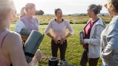 Woman standing holding stomach to represent pelvic floor issues, among other women holding yoga mats outdoors