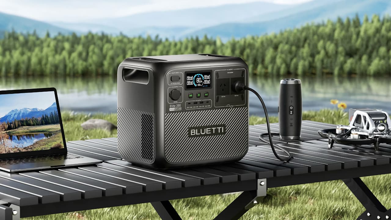 The Bluetti Elite 200 V2 on a camping table charging a drone, bluetooth speaker and laptop