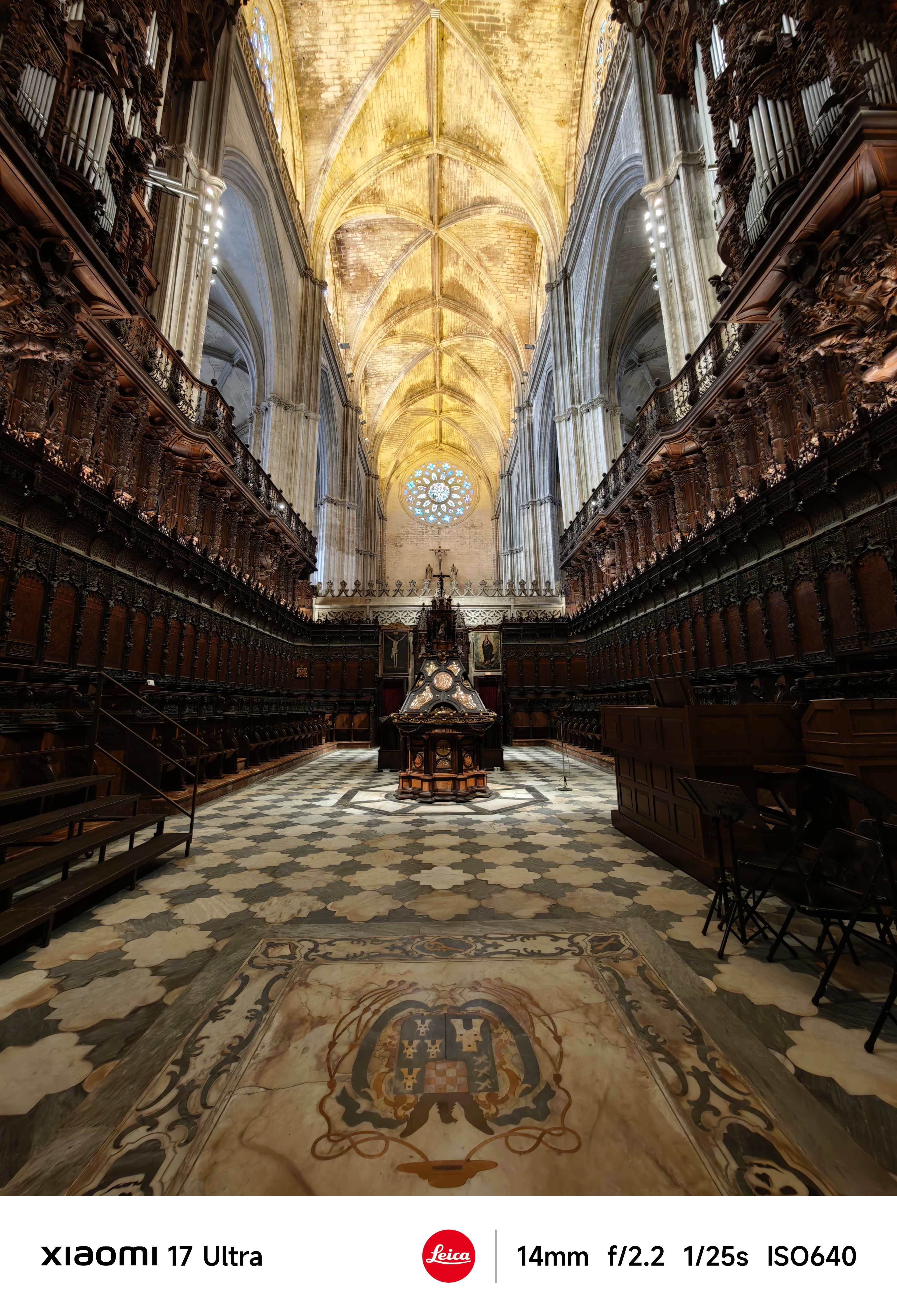 Symmetrical view inside a cathedral choir with dark carved wooden stalls, patterned tiled floor, and illuminated vaulted ceiling leading to a circular stained-glass window.