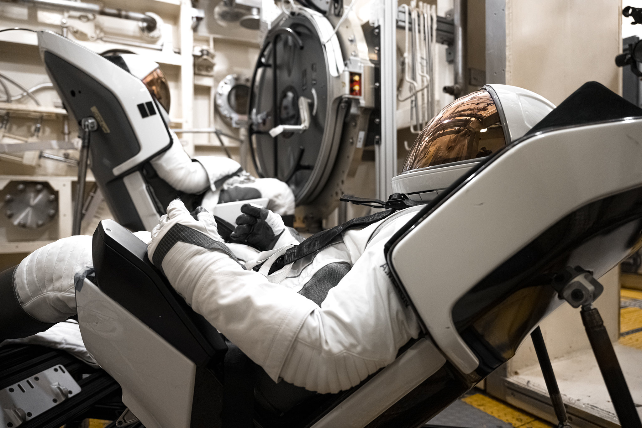 Two astronauts, one in the foreground looking to the left and the other in the background looking to the right, are wearing white spacesuits and leaning against a set of black and white buckets.