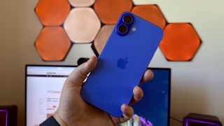 A blue iPhone 16 Plus being held up in front of orange wall lighting.