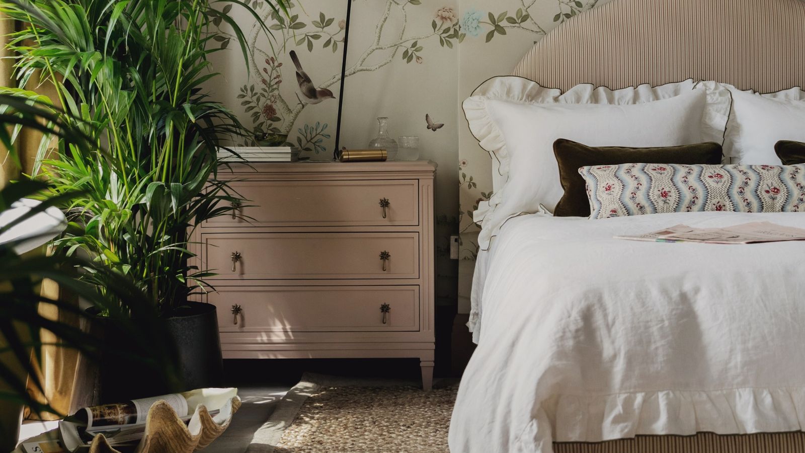 romantic pale pink bedroom with floral mural wallpaper, a pink striped headboard, white ruffle trim bed linen and large indoor plants