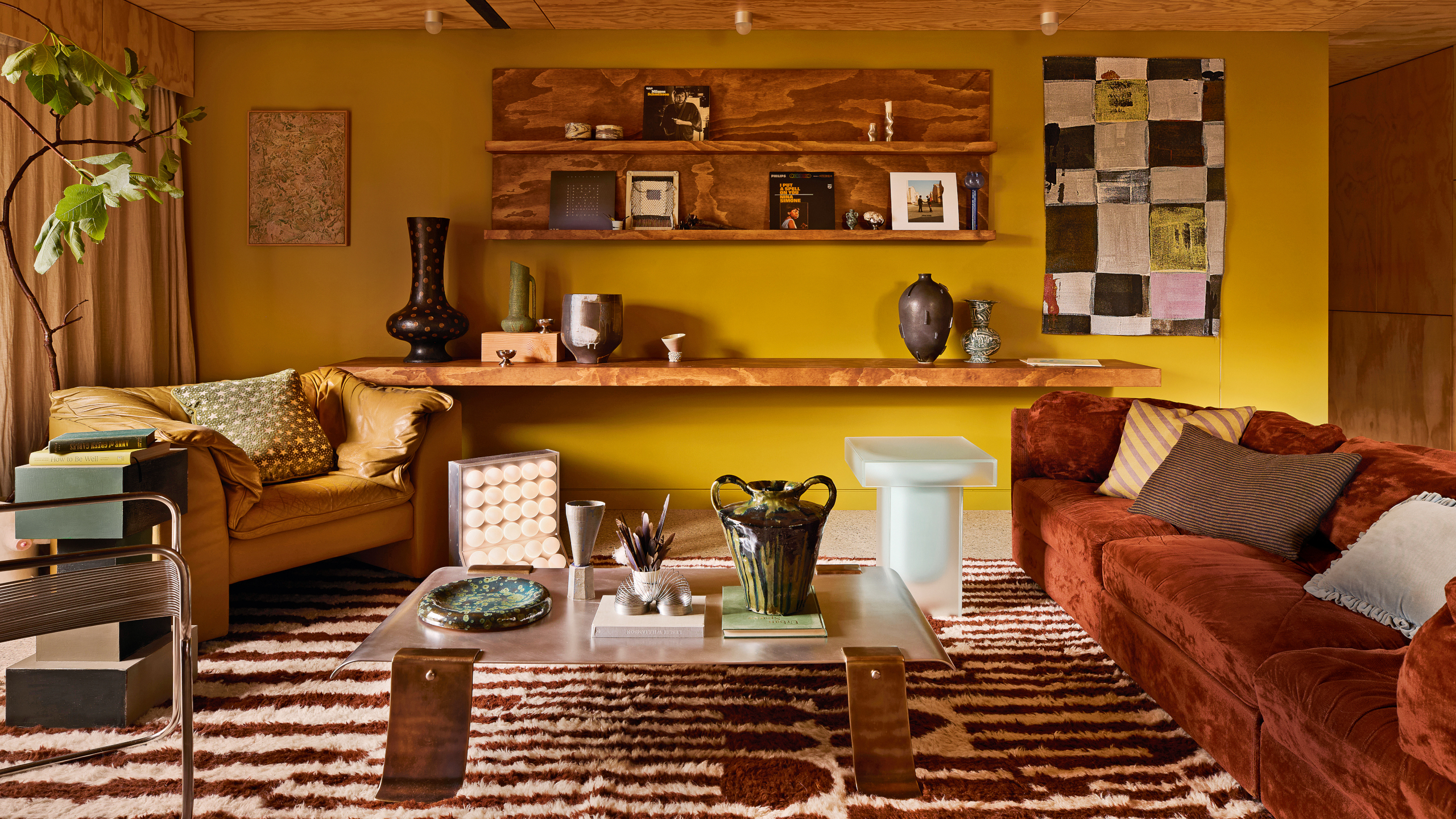 Living room with ochre walls, russet-coloured velvet sofa and red-tan leather sofa, almond-shaped stone coffee table and dark blue low-level cabinetry, with a brown and white striped rug