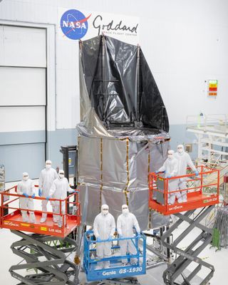 Seven technicians in white "bunny suits" stand in front of a large space telescope inside a white-walled clean room