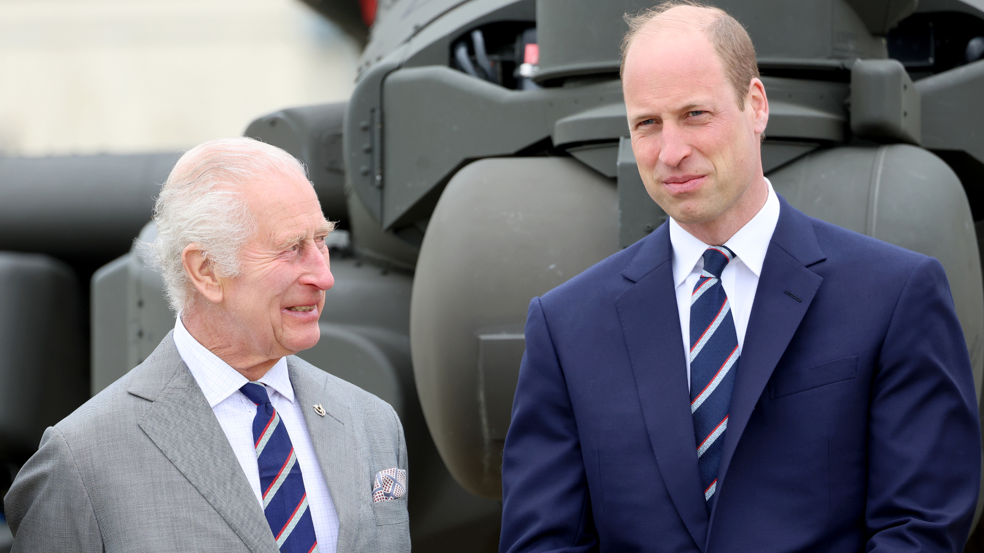 King Charles wears a gray suit and smiles at son Prince William, who has a bald head and is wearing a navy suit