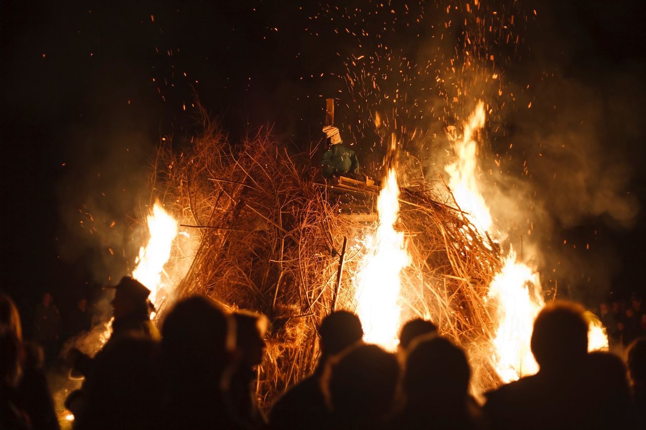 A large bonfire blazing at night with sparks flying into the air, watched by a crowd of silhouetted people; a Guy effigy can be seen on top of the burning pile.