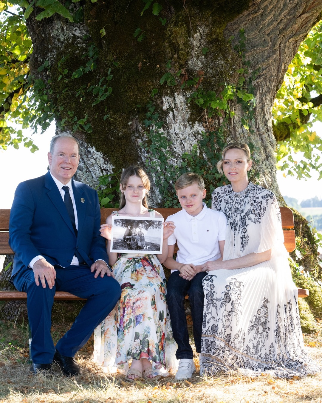Prince Albert, Princess Charlene and their twins sitting in front of a tree holding a black and white photo