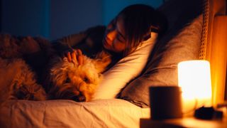 A woman with dark hair cuddles her dog in bed next to a calming orange bedside lamp